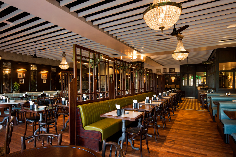 Interior of Dishoom restaurant with wooden tables, green leather banquettes, dark wood panelling and chandeliers overhead.