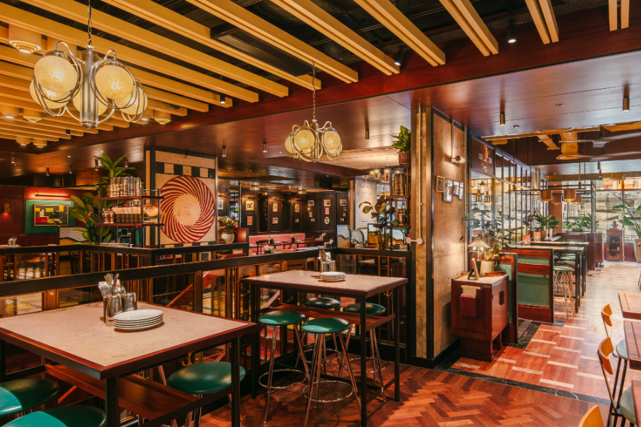 Interior of Dishoom Glasgow with high tables, green stools, warm wood panelling and decorative lighting.