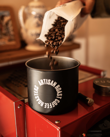 Artisan Roast coffee beans being poured into a black coffee container beside a red coffee roaster.