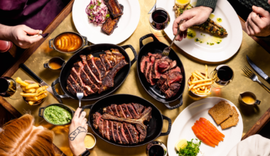 Overhead view of a Hawksmoor table in Edinburgh filled with large sharing steaks, fries, sauces, grilled vegetables, smoked salmon and red wine, with diners gathered around the table carving and serving food