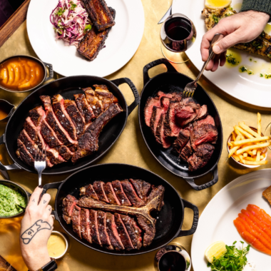 Overhead view of a Hawksmoor table in Edinburgh filled with large sharing steaks, fries, sauces, grilled vegetables, smoked salmon and red wine, with diners gathered around the table carving and serving food