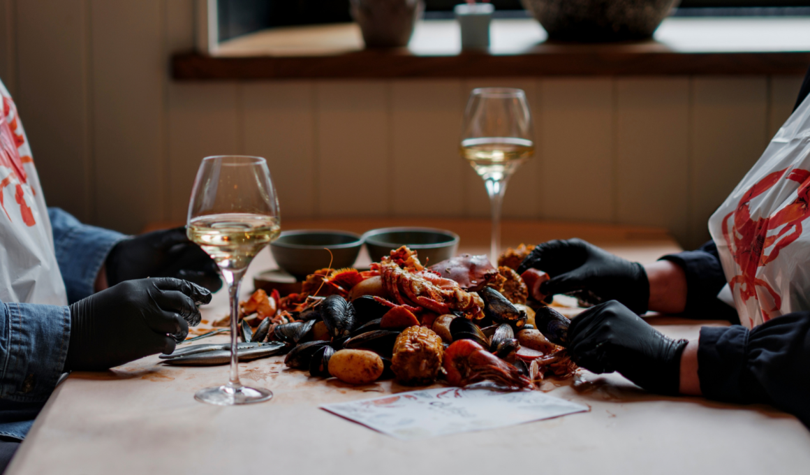 Two diners wearing lobster bibs and black gloves sharing a seafood boil with Scottish shellfish and glasses of white wine at Dulse Leith.